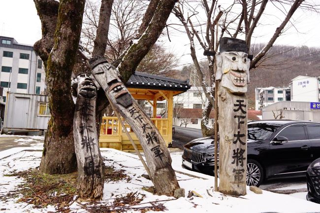 큰 길가에 천하대장군 셋, 왼쪽 둘은 쓰러져 뒤의 느티나무에 기대어 있음, 뒤로 보이는 건물들, 우측에 주차된 검은 승용차 1,