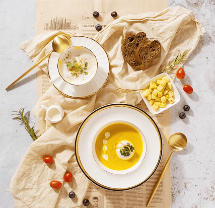식탁에 노란 호박 스프가 담긴 그릇과 양송이 스프가 담긴 그릇이 있다. 그릇 옆에는 금색 숟가락이 하나씩 놓여있고, 갈색 통밀빵과 방툴토마토가 있다.