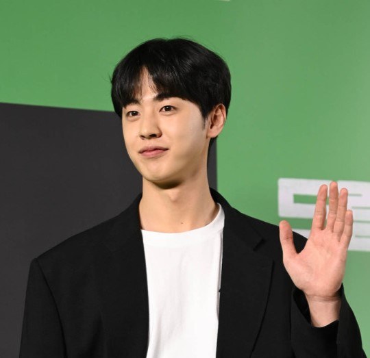 검은 재킷에 흰 티셔츠 차림으로 초록색 배경 앞에서 손을 흔드는 배우 김현진