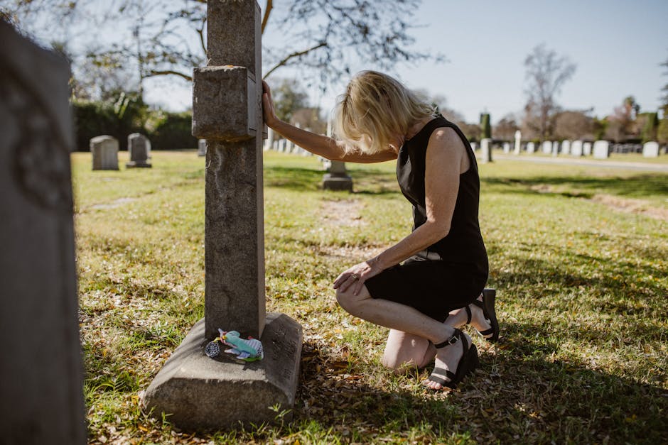 A woman in a black dress kneeling at a tombstone, expressing grief in a sunlit cemetery.