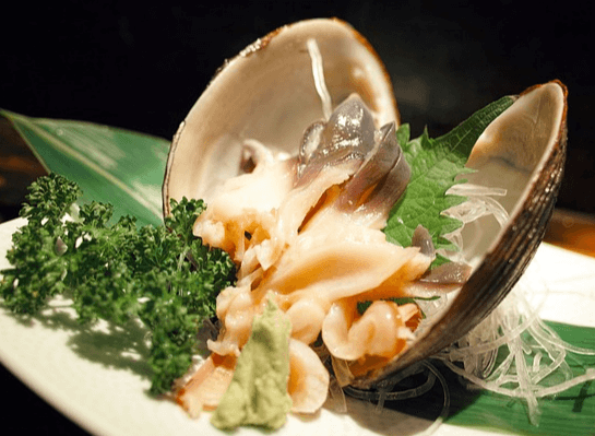 네모난 하얀 접시 위에 커다란 조개구이 한 마리가 있고 앞에는 초록색 파슬리가 있다. 조개껍질은 벌려졌고 하얀 속살이 보인다.