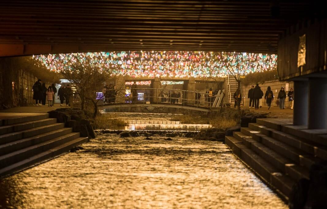 청계천 다리 아래를 가득 채운 화려한 등불이 밤하늘처럼 빛나는 장면과 주변을 산책하는 사람들의 모습