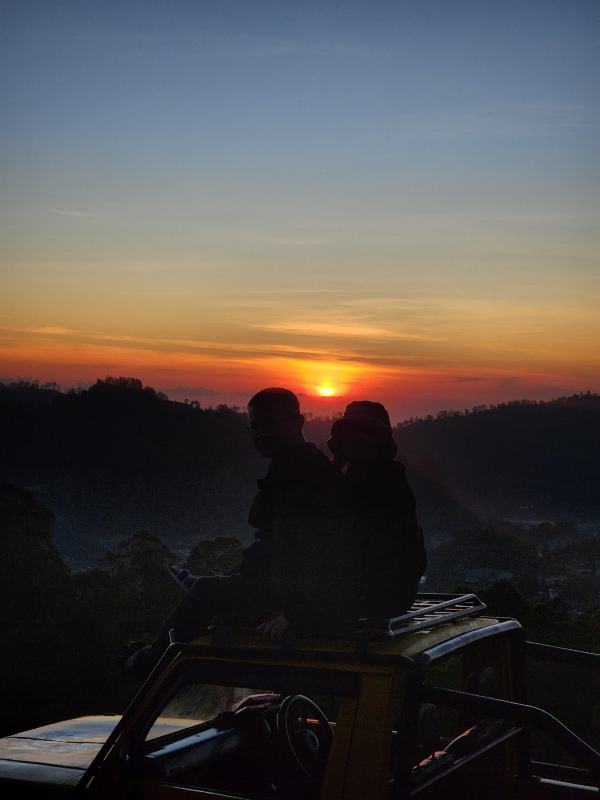 구름 사이로 노란색 태양이 떠오르고 있다. 주변은 어둡다. 두 사람의 그림자도 보인다.