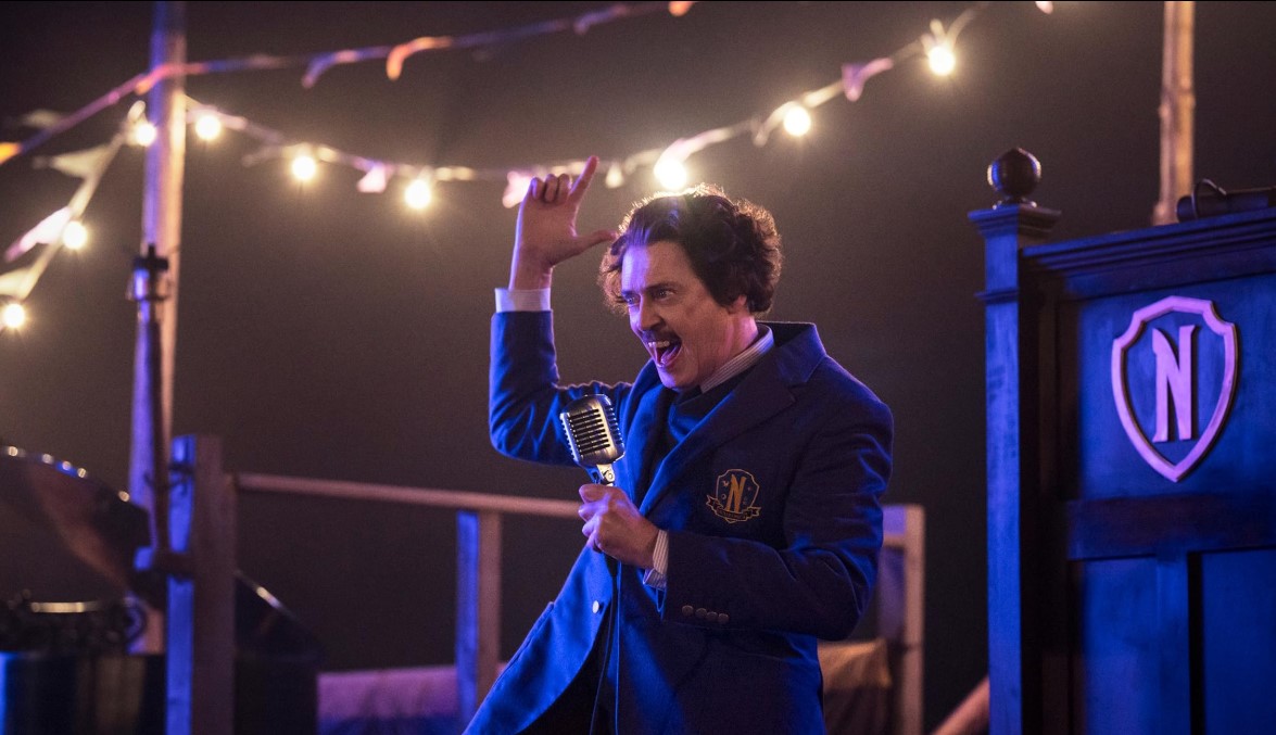 넷플릭스 웬즈데이 시즌2 신규 캐릭터 배리 도트 교수 역의 스티브 부세미 &ndash; 네버모어 아카데미 교복을 입고 무대에서 마이크를 들고 있는 장면