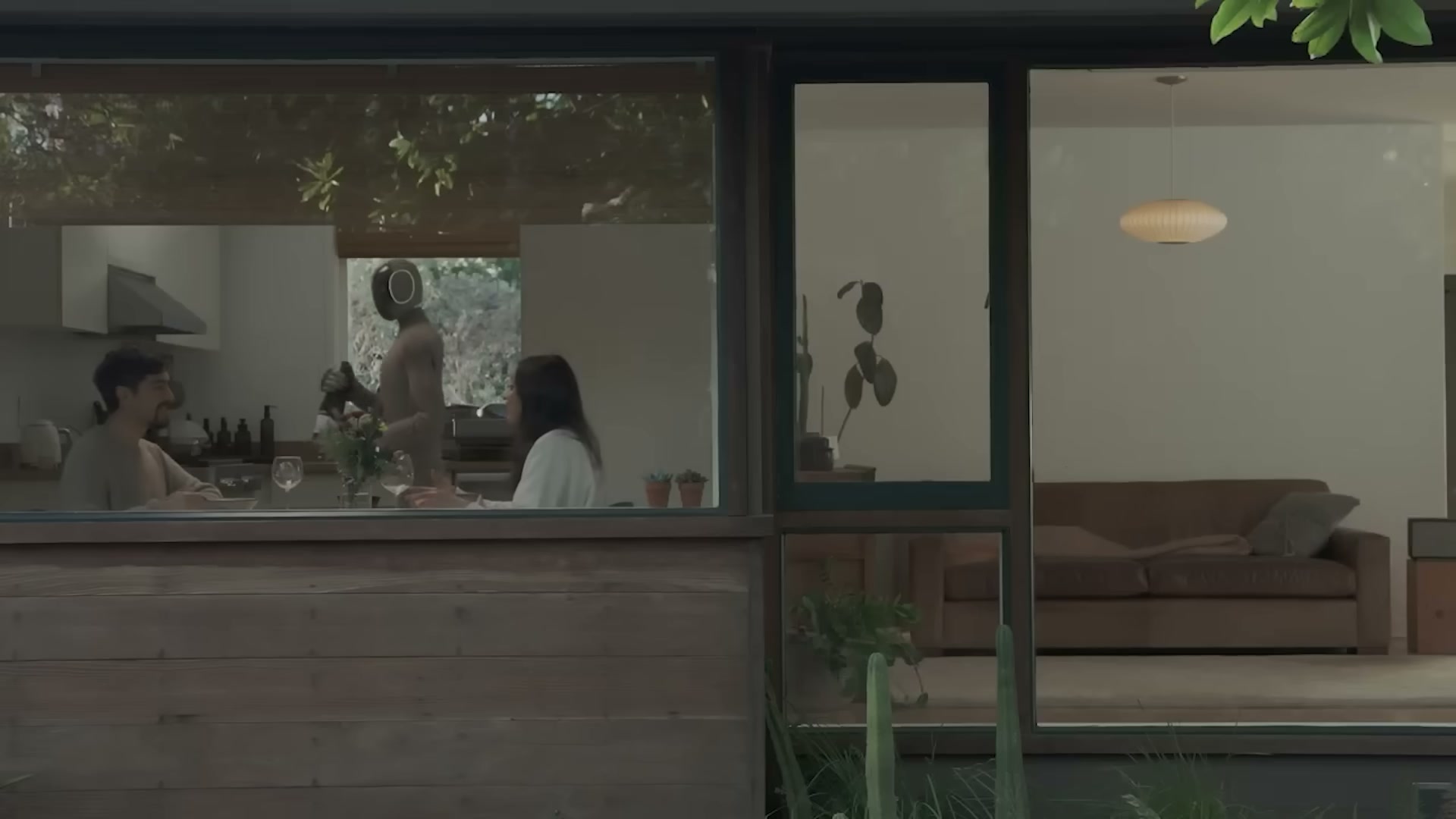 넓은 창과 거실이 보이는 집의 부엌에서 회색 휴머노이드 로봇이 한 부부 옆에서 와인 잔을 가져다 놓고 있다.