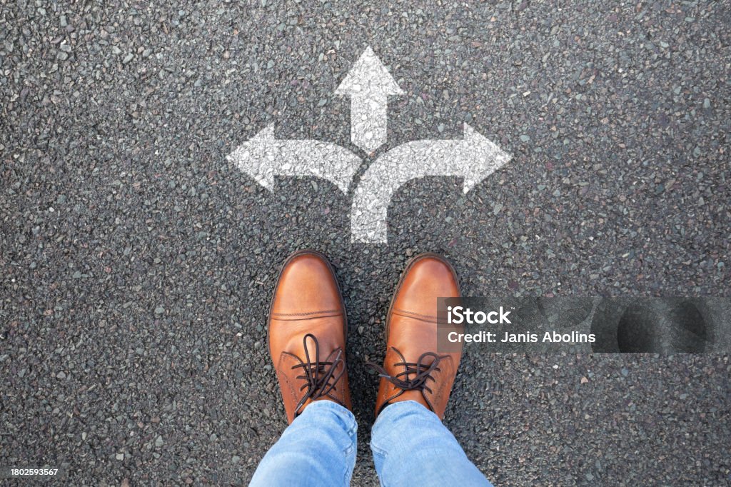 갈림길 바닥에 서 있는 사람의 신발 사진. 여러 방향으로 나뉜 화살표가 보인다. 선택의 갈림길을 의미한다.