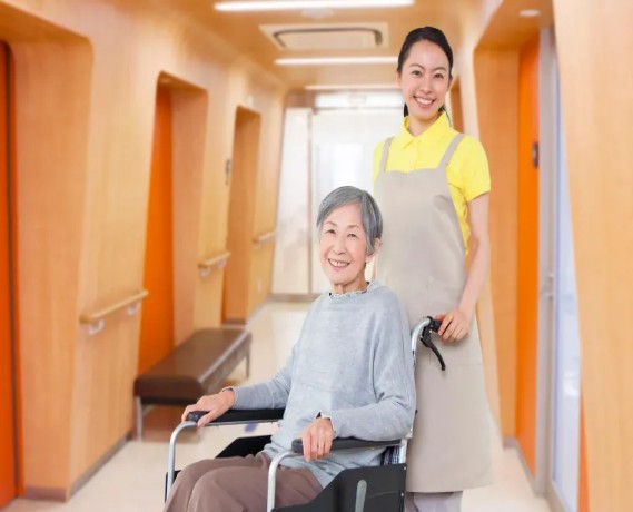 밝은 병동 복도에서 휠체어에 탄 환우님을 미소 지으며 밀어드리는 요양보호사의 따뜻한 동행