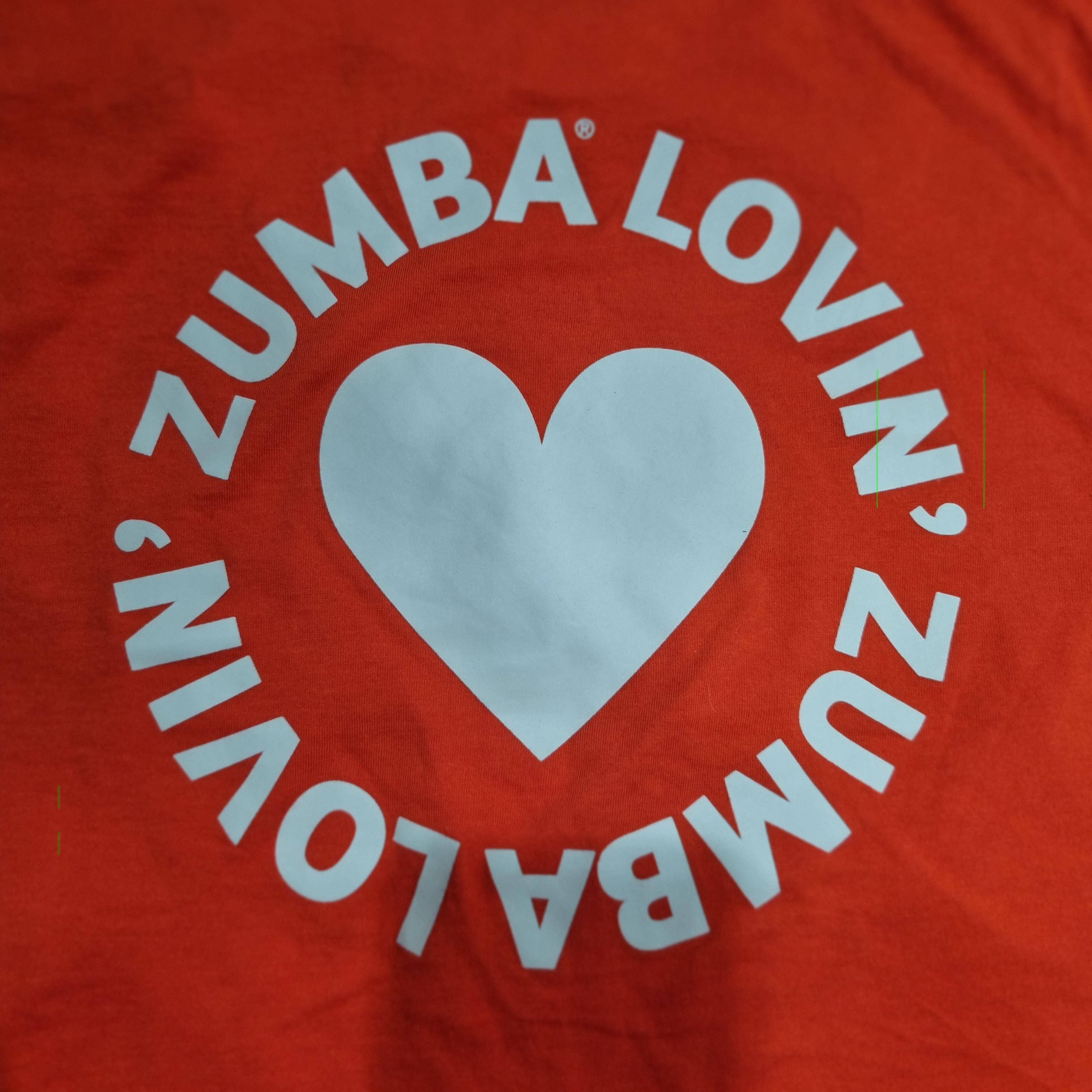 "빨간 배경의 티셔츠 중앙에 흰색 하트 아이콘이 있고, 그 주변을 따라 'ZUMBA LOVIN''이라는 문구가 원형으로 반복되어 인쇄되어 있는 디자인