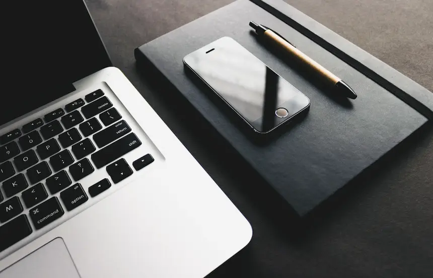 노트북 옆에 다이어리 그 위에 핸드폰 펜이 놓여있는 사진