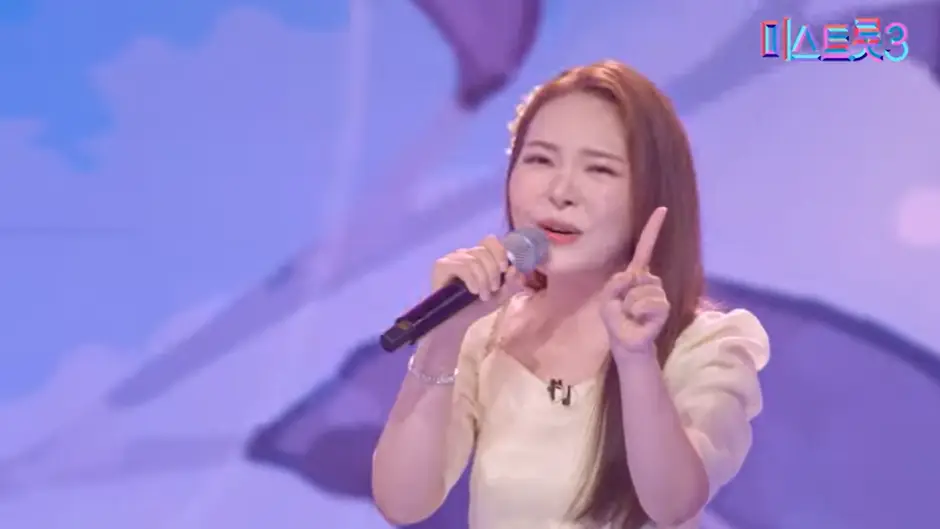 미스트롯3- 보라색 배경 노란 드레스를 입고 긴머리를 늘어뜨린채 오른손으로 마이크를 잡고 노래하는 미스김