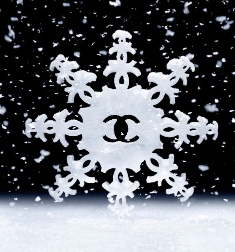 image shows a snowflake design with the Chanel logo (&quot;CC&quot;) in the center. The snowflake appears to be stylized with intricate details and is surrounded by falling snowflakes, creating a festive, wintry atmosphere. The design highlights Chanel’s branding, integrating the iconic logo within the snowflake structure for an elegant seasonal theme.