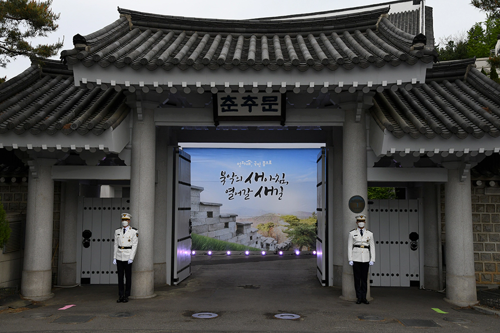 기와 지분느오 이루어진 춘춘문이라는 하얀 대문이 열려있고 그 양옆에 하얀 제복을 입고 마스크를 쓴 두 사람이 서있다.