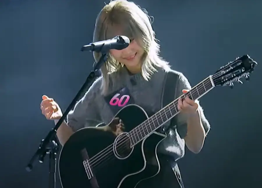 싱어게인 3- 검은색을 배경으로 가슴에 60번호표를 달고 단발머리를 늘어뜨린채 웃으며 기타를 치는 60호 상반신