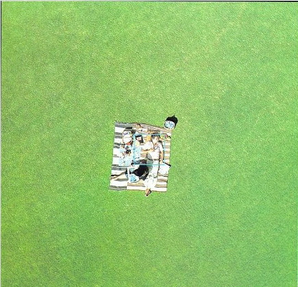 지상 10미터 상공에서 내려다본 장면. 넓은 초록 잔디밭 가운데 줄무늬 담요가 펼쳐져 있고, 그 위에는 한 남성이 누워 있고 그의 오른쪽 다리 곁에는 아내가 다리를 쭉 뻗은 채 책을 읽고 있다. 담요 위에는 소풍 음식과 소지품이 가지런히 놓여 있으며, 두 사람은 고요하고 따뜻한 한낮의 여유를 즐기고 있다.