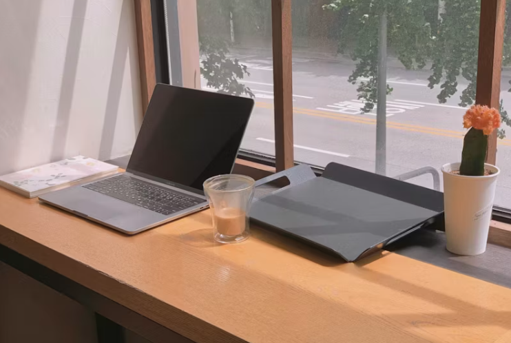 햇살이 비치는 창가 책상 위에 닫힌 노트북과 커피잔, 화분이 놓여 있으며, 창밖으로 나무와 도로가 보이는 모습