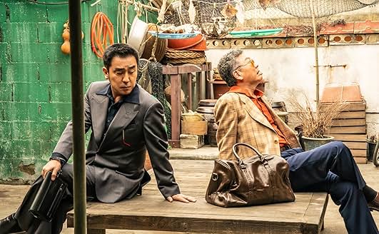 야외 평상 위에 앉아 서로 등을 돌린 두 명의 남성&amp;#44; 한 명은 정장을 입고 서류가방을 들고 있음