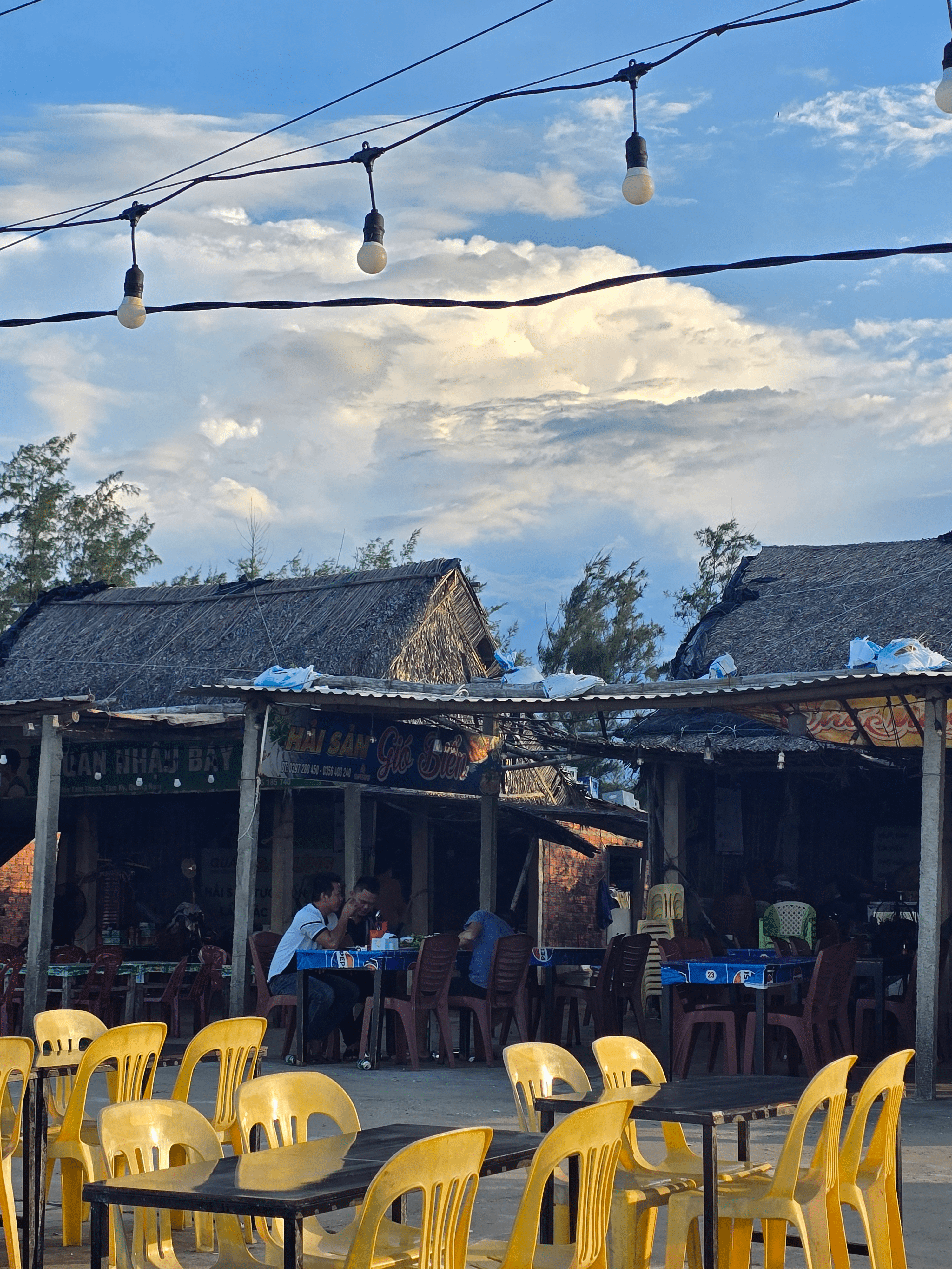 베트남 다낭 땀끼 해변가의 현지 음식점 풍경. 노란 플라스틱 의자가 놓여 있고, 사람들이 식사하며 담소를 나누고 있다. 전구가 매달린 하늘은 푸르고 구름이 흘러가며 여유로운 분위기를 자아낸다.