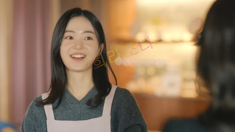 실내에서 한봄(김승윤)이 밝게 웃으며 대화를 나누고 있다. 그녀는 짙은 회색 니트 상의 위에 연보라색 앞치마를 두르고 있으며&amp;#44; 검은 머리를 단정하게 내렸다. 그녀 앞에는 다른 사람이 서 있는 듯하지만&amp;#44; 보이지 않는다.
