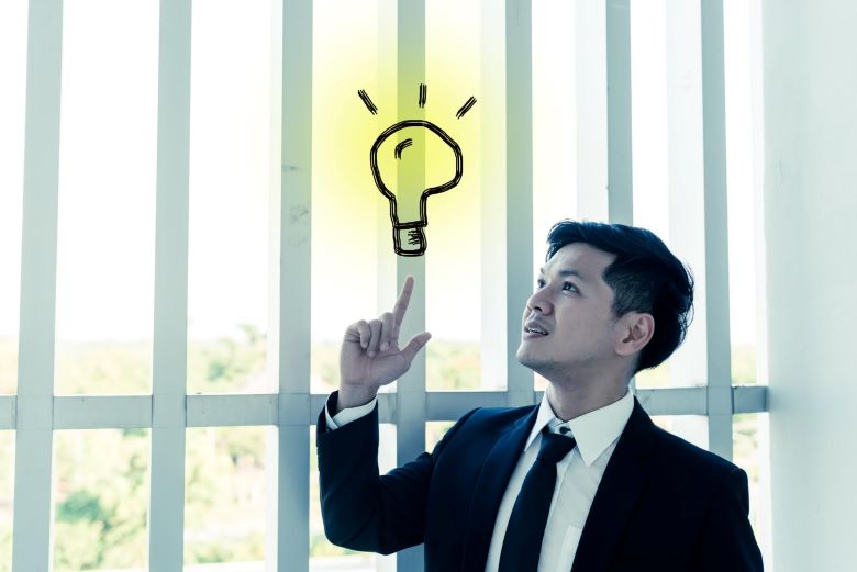 밝은 창문 앞에서 검은 정장과 넥타이를 입은 아시아 남성이 위를 보며 손가락으로 가리키고 있고, 머리 위에 노란 빛나는 전구 아이디어 아이콘이 그려진 창의적 아이디어와 영감 개념 이미지 via freepik