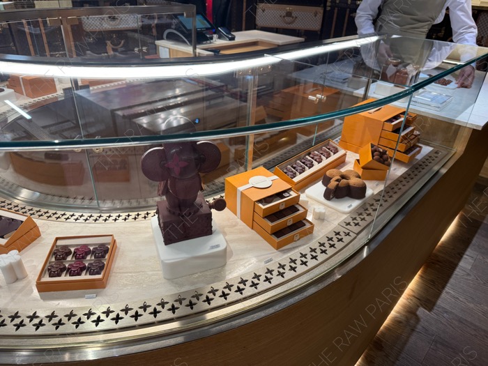 A glass pastry display case at Louis Vuitton Cafe by Maxime Frederic inside LV DREAM filled with monogrammed chocolates and cakes ❘ Une vitrine de pâtisserie en verre au Louis Vuitton Café par Maxime Frédéric chez LV DREAM avec des chocolats monogrammes