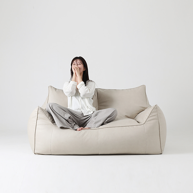 흰 배경 앞에서 흰 상의와 회색 바지를 입은 여성이 플래지어 톰빈백 2인용 아이보리 기능성 가죽 빈백 소파 가운데에 양반다리로 앉아 두 손으로 입을 가리고 웃는 모습