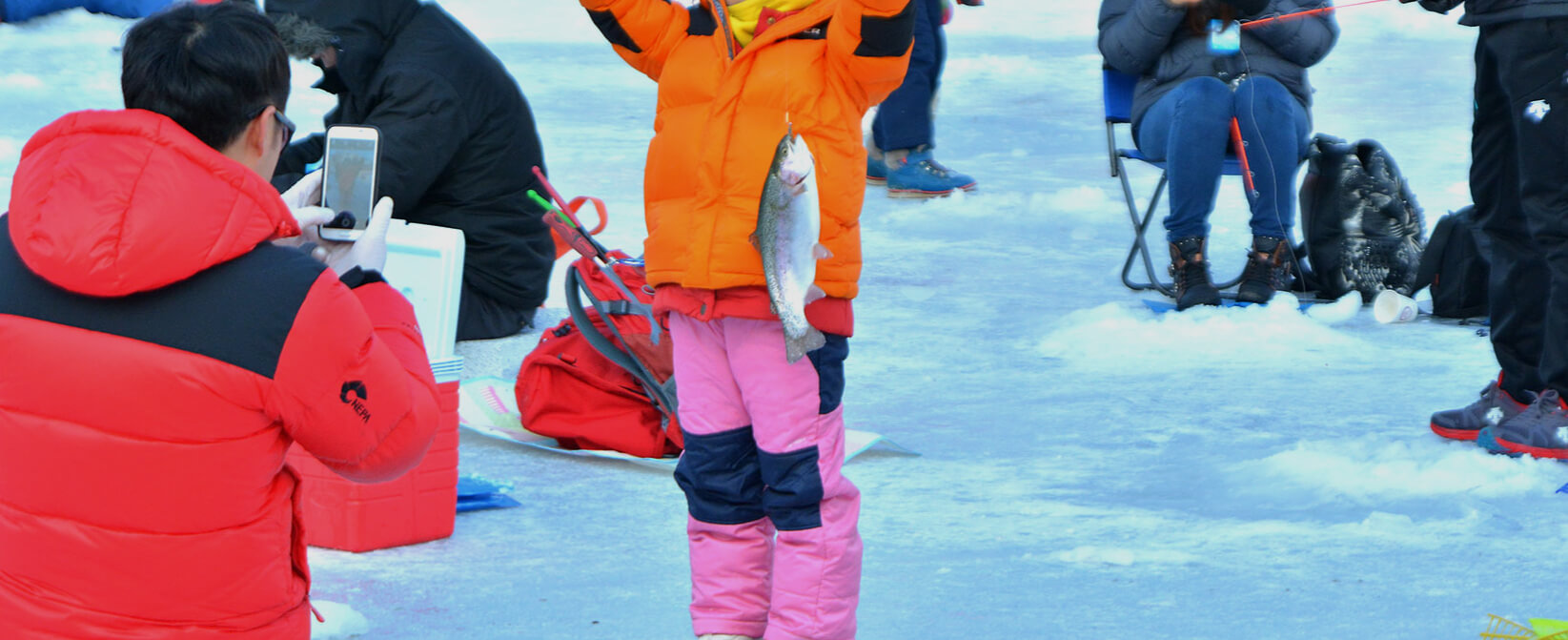 한 아이가 얼음 낚시로 잡은 송어를 들고 자랑스럽게 포즈를 취하며, 주변에 겨울 의상을 입은 사람들이 축제를 즐기는 모습