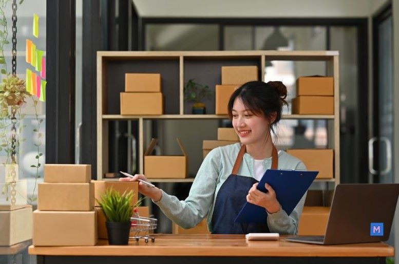소상공인 사업자가 사무실에서 배송 박스들과 함께 클립보드를 들고 미소짓는 모습, 새출발기금 신청 대상 소상공인 이미지 via canva