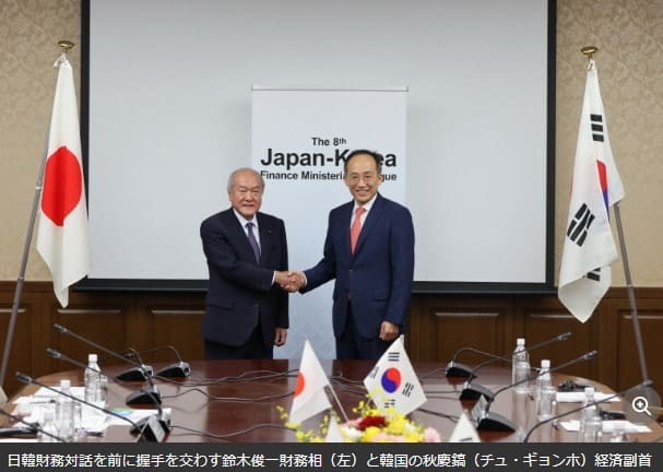 재팬 코리아라고 적혀있는 이젤 앞에서 두 남자가 정장을 입고 악수를 하고 있다.