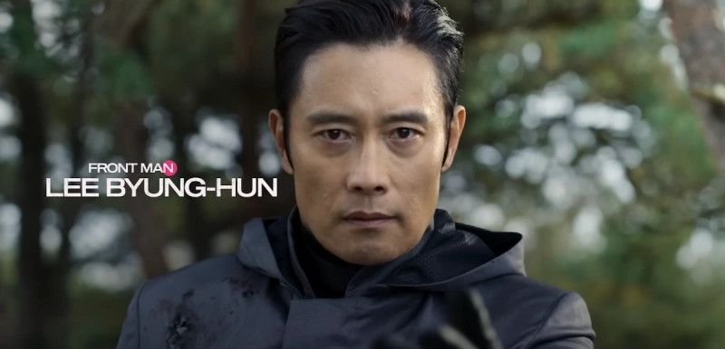 숲을 배경으로 검은 옷을 입고 어깨에 총상을 입은 프론트맨역의 배우 이병헌