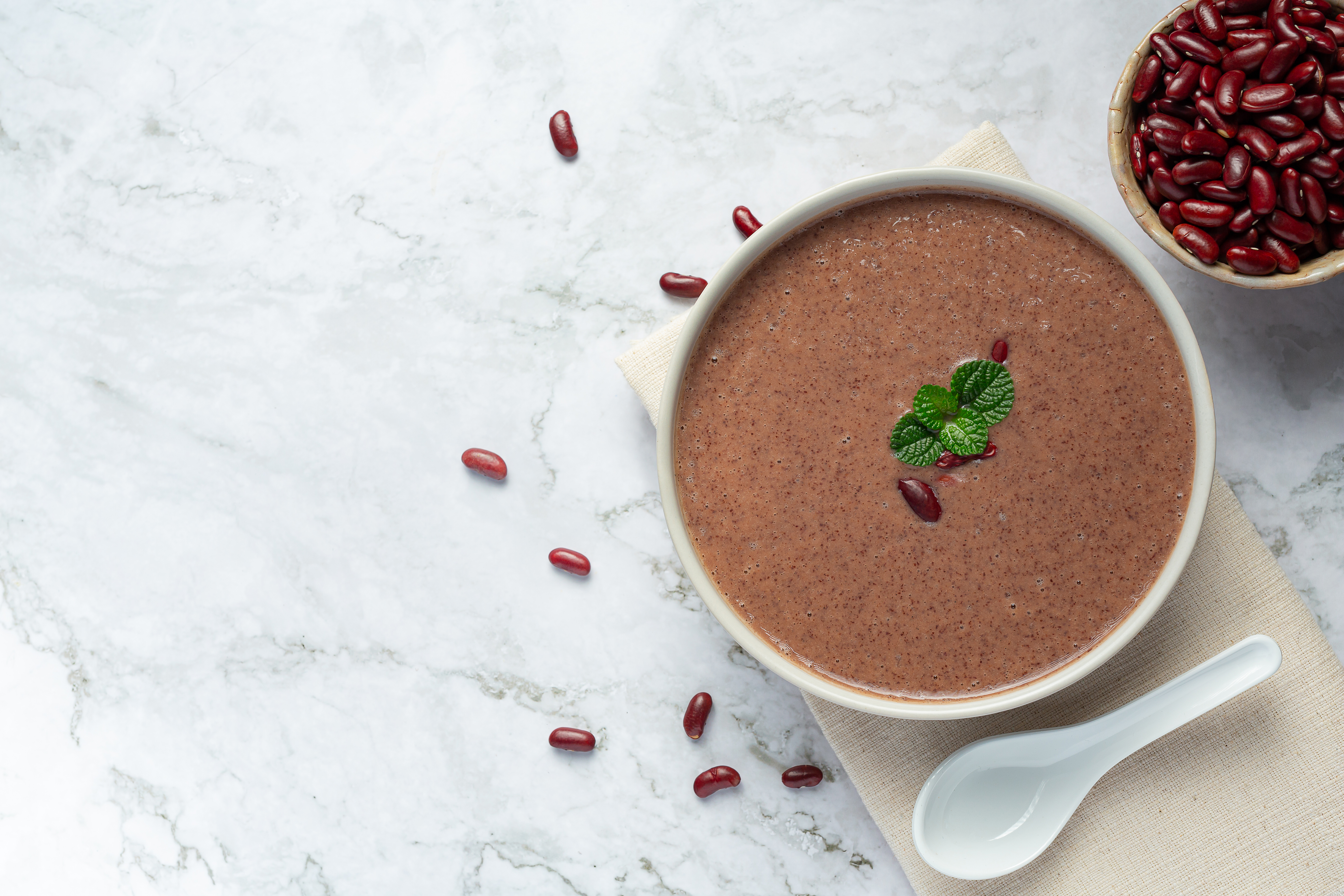 깨끗한 테이블 위에 헝겊 깔판을 깔고 그 위에 흰 그릇에 팥죽을 가득 담은 후에 팥과 잎으로 장식을 하고&#44; 그 주변에 팥을 가득 담은 그릇과 바닥에 팥을 흩어 놓은 사진