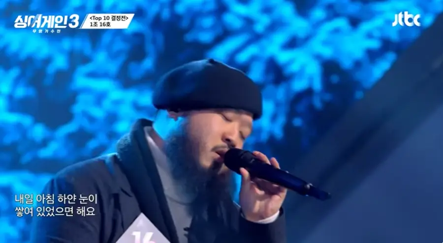 싱어게인3-
파란배경 검은색 외투에 회색 폴라티 검은 배레모에 검은 마이크를 들고 노래하는16호의 오른쪽 모습