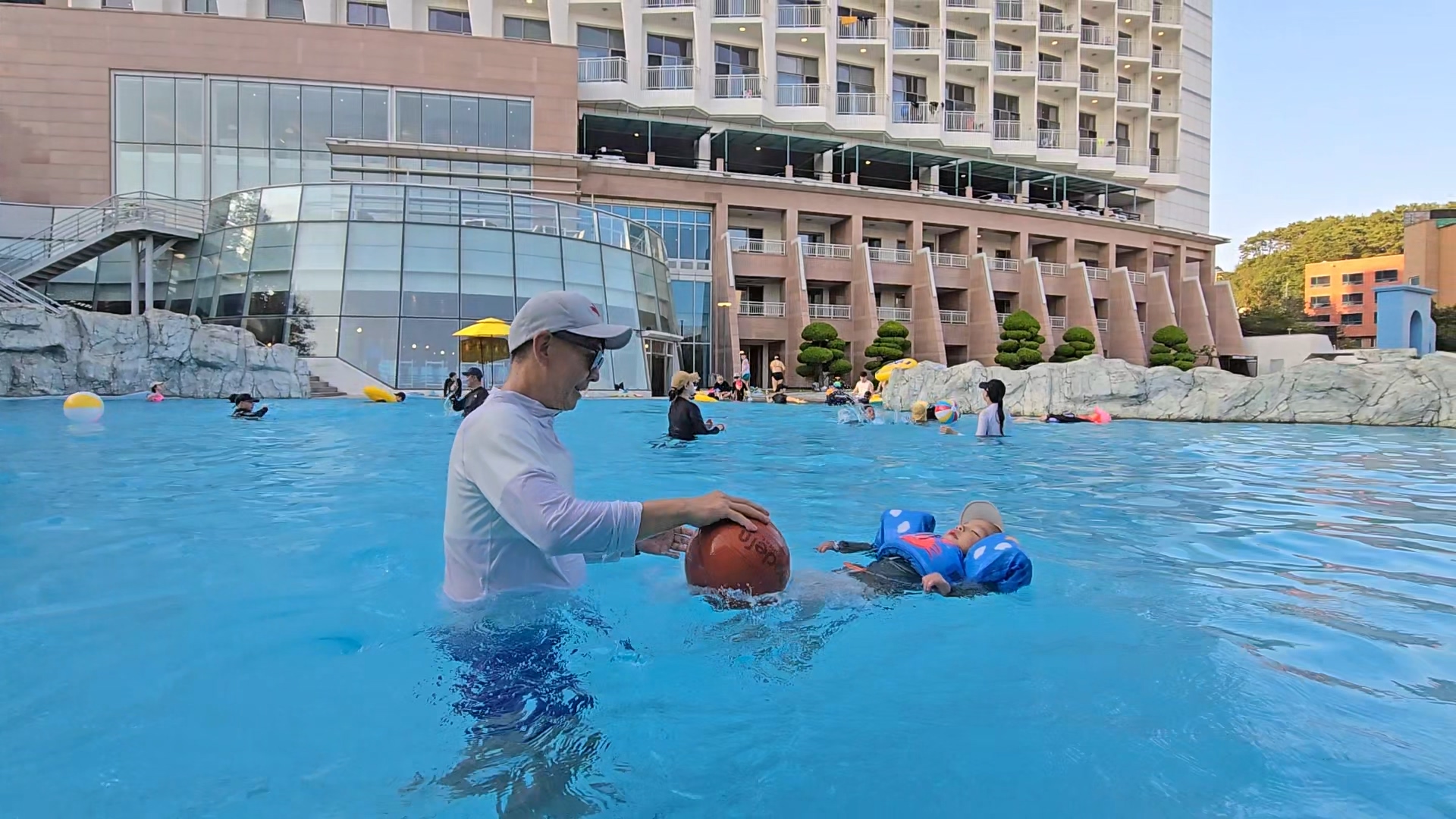 더위크앤 아쿠아벤처 워터파크 야외 수영장에서 아빠와 아들이 함께 물놀이를 즐기고 있는 모습. 아이는 구명조끼를 착용한 채 물 위에 누워 있고, 아빠는 공을 잡고 아이를 바라보며 웃고 있다. 뒤로는 리조트 건물이 보이며, 여러 가족이 물놀이 중이다.