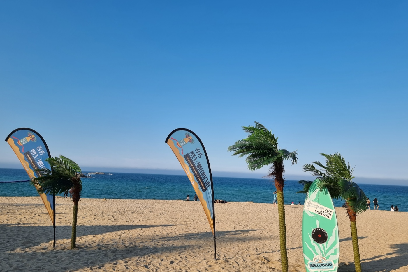 경포해변 주변에 홍보조형물(배너광고)과 인조 야자수, 써핑 보드 등이 있고 사람들은 해변가에서 바다를 바라보고 즐기고 있는 모습이다.