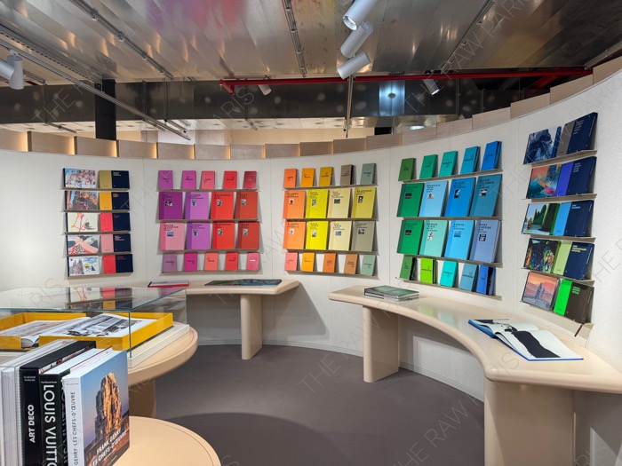 A curved display wall at LV DREAM store in Paris filled with colorful Louis Vuitton books and travel guides ❘ Un mur d'affichage incurvé chez LV DREAM à Paris rempli de livres Louis Vuitton colorés et de guides de voyage