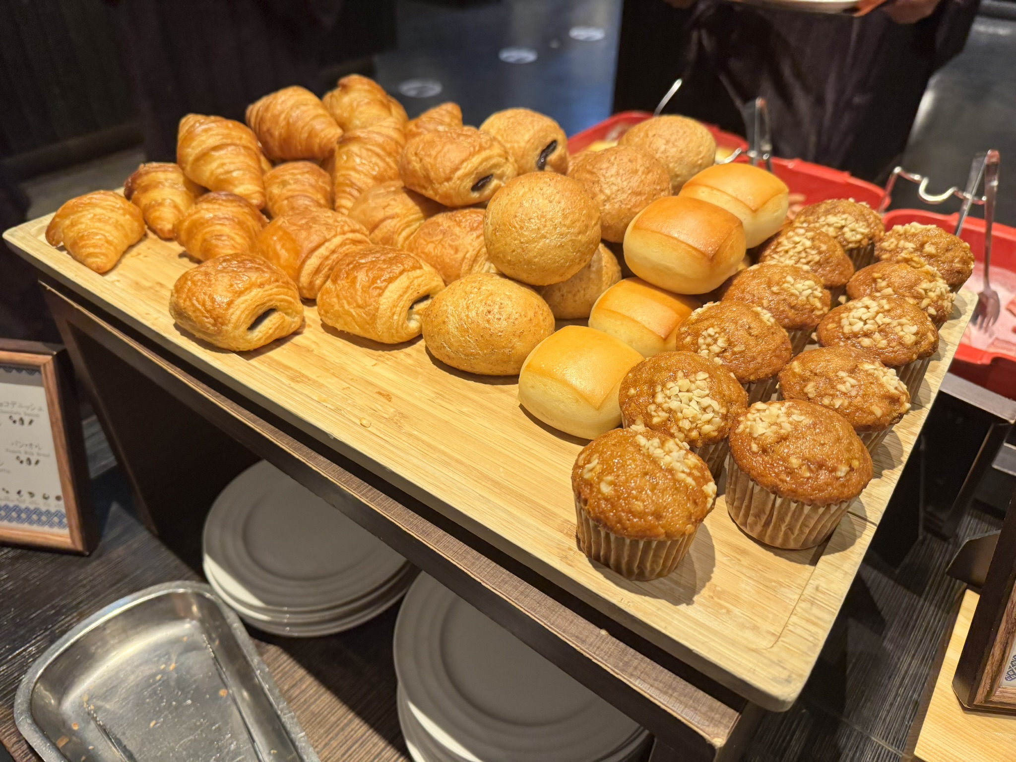 나무 쟁반 위에 가득 놓인 조식용 빵들. 노릇하게 구워진 크루아상, 뺑 오 쇼콜라, 모닝빵, 그리고 고소한 견과류가 뿌려진 머핀들이 먹음직스럽게 진열된 모습.