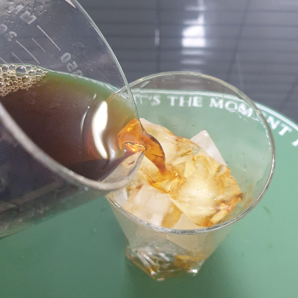 매트위에 얼음이 담긴 유리컵이 있다. 이 유리컵에 추출된 커피를 붓고 있다.