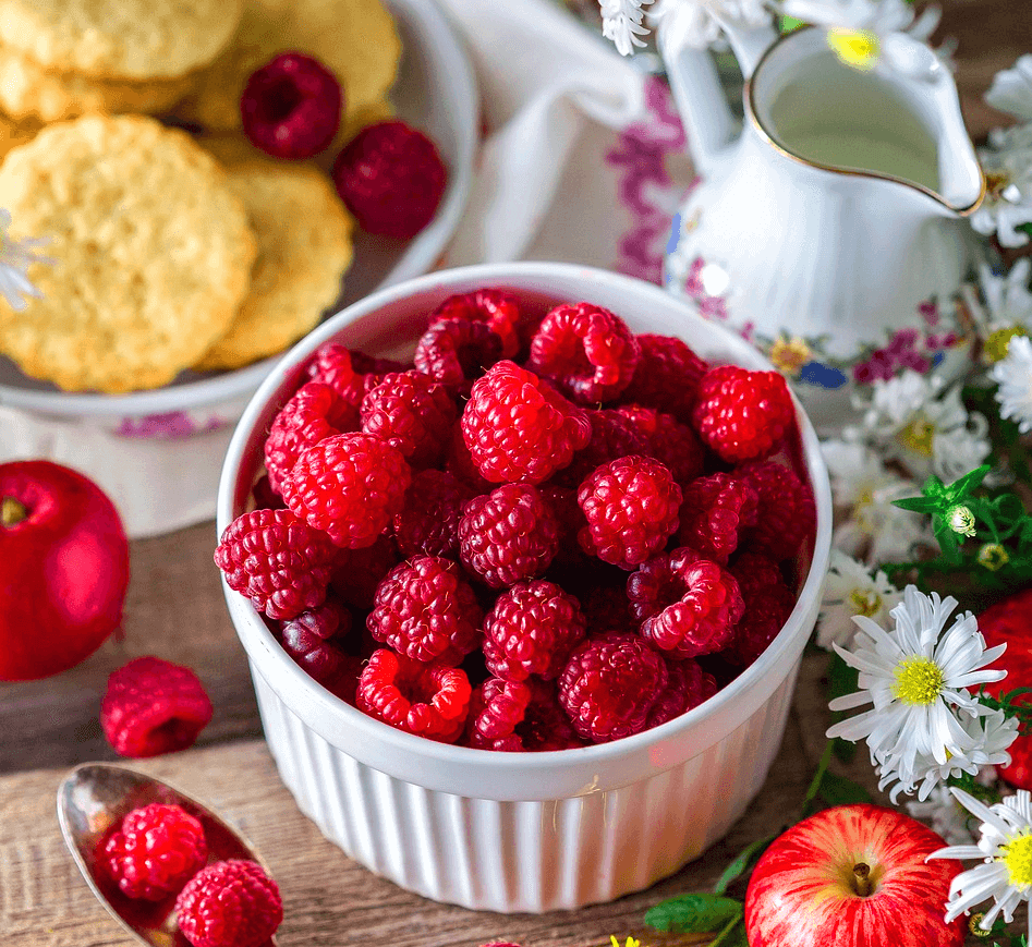 하얀 그릇에 붉은 라즈베리들이 가득하다. 주변엔 둥그런 비스킷이 담긴 접시가 있고 하얀 꽃들도 있다.