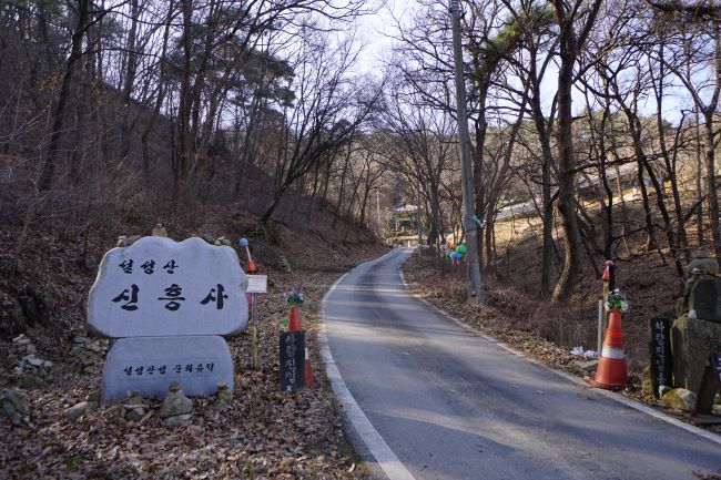 신흥사 사찰 전경, 왼쪽에 흰 대리석에 새긴 신흥사 절 이름 검은 글씨, 절로 올라가는 포장도로, 전경을 가리고 있는 키 큰 나무들,