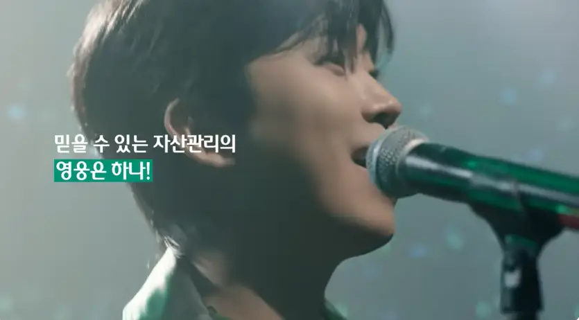 임영웅- 검은 무대 불빛들이 반짝이는 콘서트 장 중앙 임영웅 얼굴 앞 마이크가 크게 잡힌 장면 좌측 작은 흰글씨 믿을 수 있는 자산관리의 초록네모 안 흰글씨 영웅은 하나!