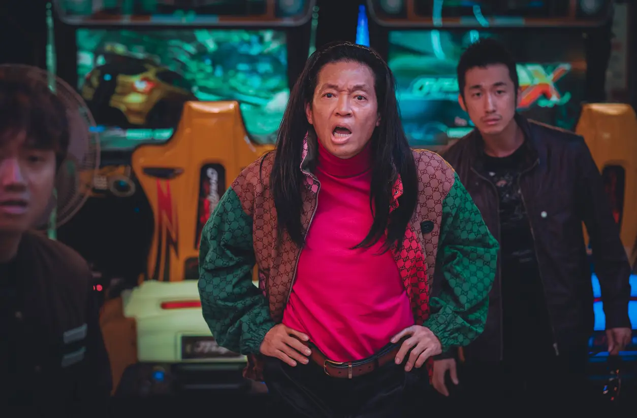 범죄도시-붉은색과 초록색이 섞인 점퍼를 입고 빨간 폴라티를 입은채 허리춤에 두팔을 얹고 화내는 장이수