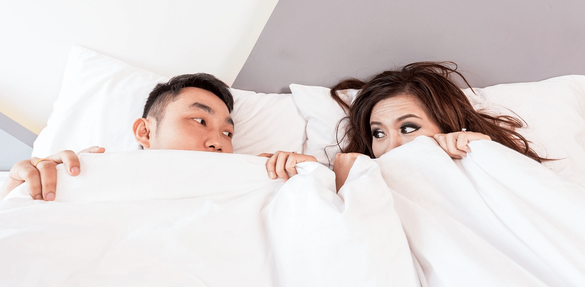 침대 위에 누워 이불을 덮고 서로 쳐다보고 있는 남성과 여성