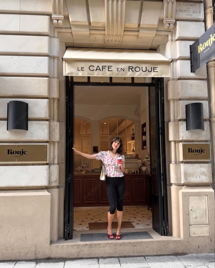 ⁠Jeanne Damas standing at the entrance of Le Cafe en Rouje in Paris wearing a floral blouse and holding a coffee cup⁠