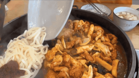 익은 닭갈비 옆으로 하얀 우동사리와 고추장 양념을 철판에 붓는 장면. 김이 모락모락 올라오며 면이 닭갈비 국물 속으로 들어가고 있다.