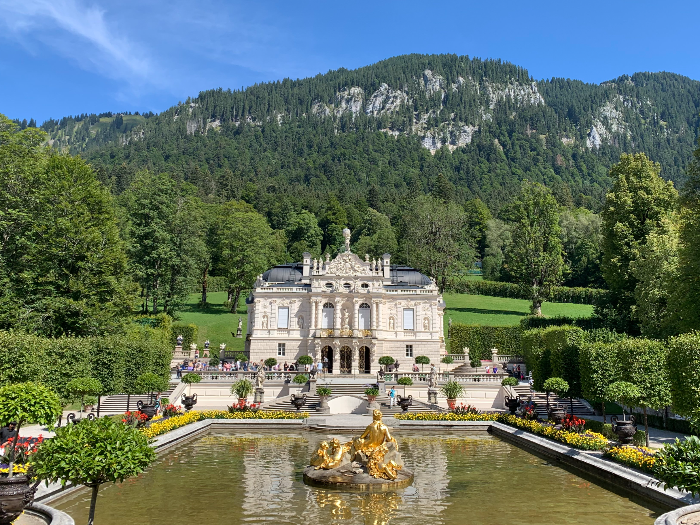좀 더 멀리서 찍은 린더호프 궁전 Linderhof Palace. 그 앞으로 금색으로 칠한 동상이 있는 호수와 궁전 뒤에는 산맥이 보인다.