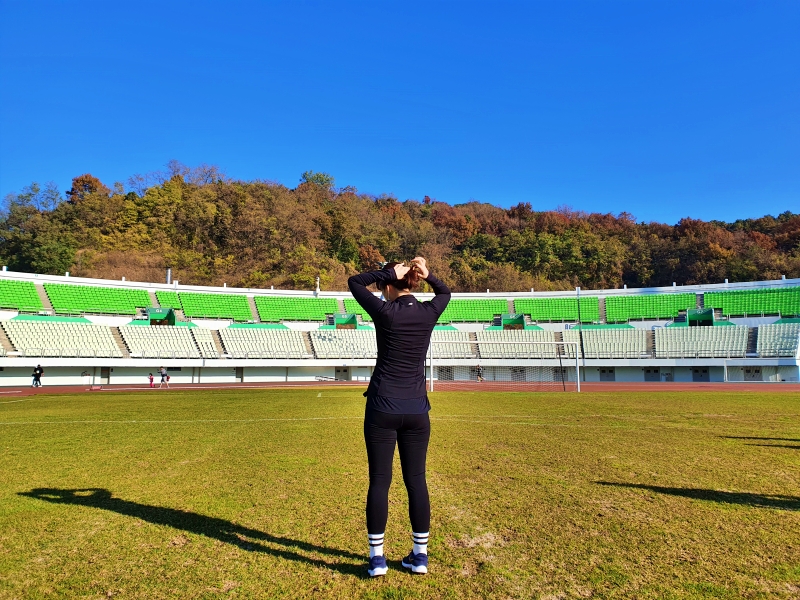 축구경기장에서 뒤로 골대가 보이고 검은색 옷을 입은 여자가 뒤로 돌아서서 머리를 묶고 있다.