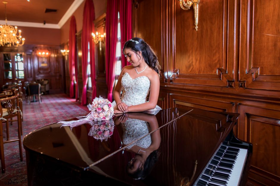 A beautiful bride in a white dress stands beside a grand piano in a luxurious room, holding a bouquet of flowers.