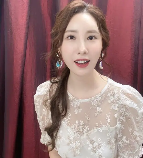 현역가왕- 빨간커튼앞 갈색 긴 웨이브머리를 아래쪽으로 하나로 묶고 망사원피스를 입고 미소짓고 있는 주미