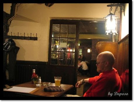 영국 런던 근교의 한 펍 내부. 나무 테이블 옆 벤치에 붉은색 상의를 입은 남성이 앉아 있고, 테이블 위에는 맥주잔과 비니, 그리고 케첩병과 양념병이 놓여 있다. 배경에는 유리문 너머로 다른 손님들이 대화를 나누고 있으며, 전체적으로 따뜻하고 조용한 분위기가 감돈다.