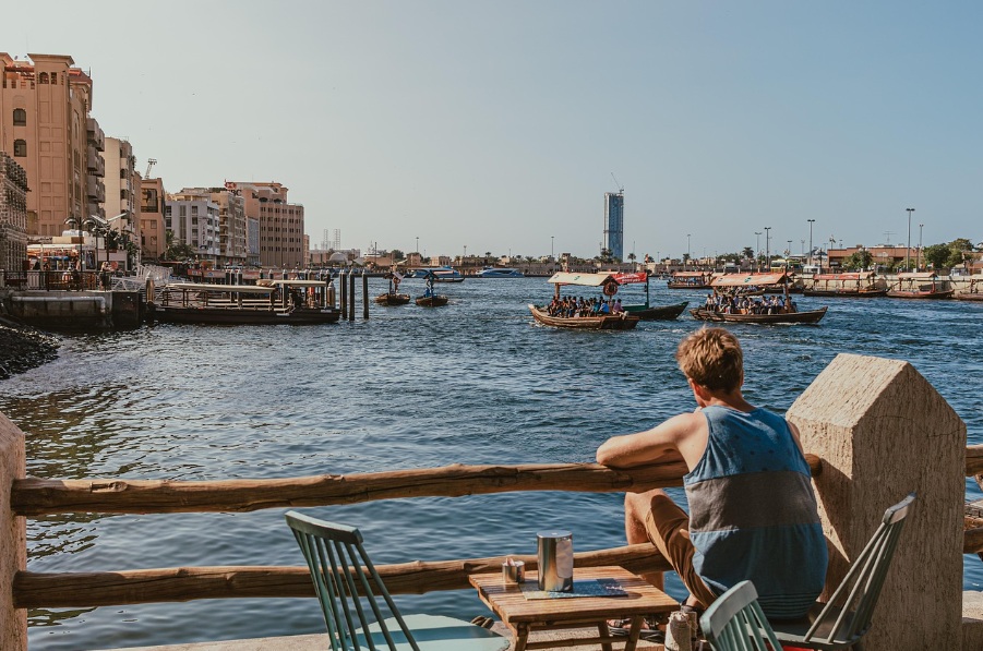 두바이 크릭(Dubai Creek)의 야외 테라스에 앉아 전통 배 아브라가 떠다니는 수변 풍경을 감상하는 여행자의 뒷모습. 멀리 현대적인 빌딩과 고즈넉한 올드타운의 건물이 어우러진 평화로운 오후 전경.