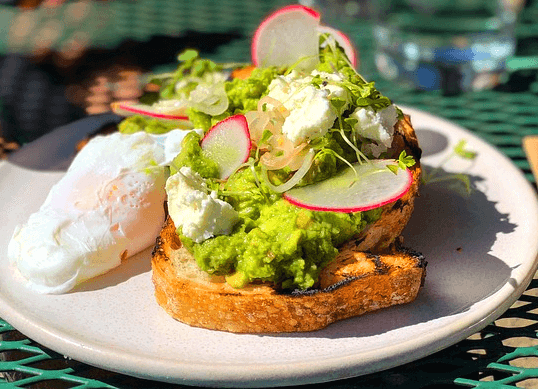 하얀 접시에 아보카도 토스트가 있다. 빵 위에 으깬 녹색 아보카도와 하얀 치즈가 있다.